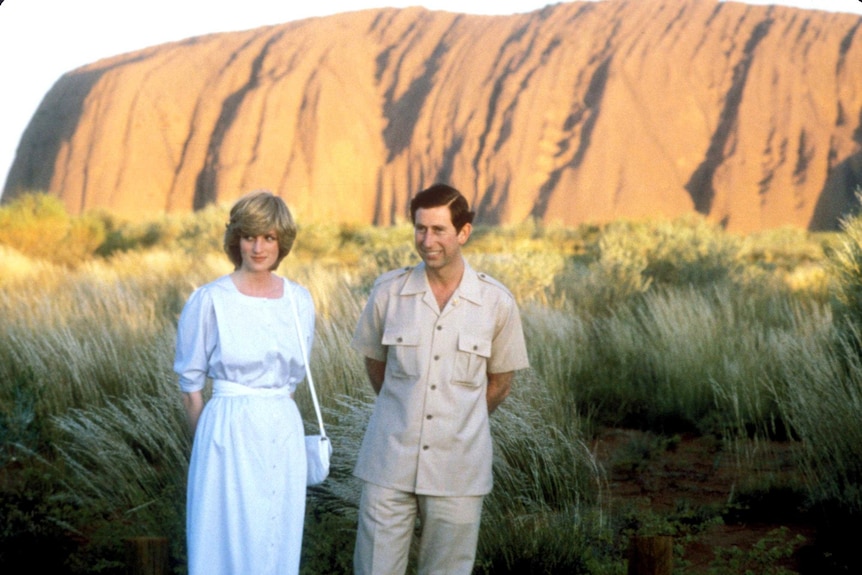 A smiling woman and man — Diana and Charles - stand  on a grassy plain at the foot of Uluru.