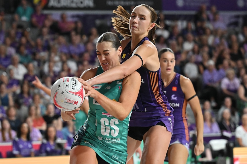 A player tries to grab the ball as another reaches over her shoulder 