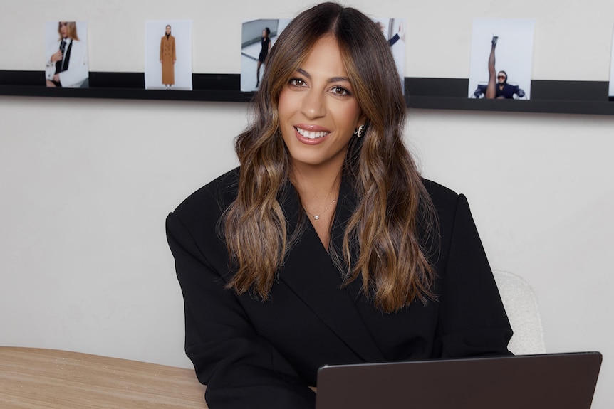 Business women in all black smiles at the camera. A laptop and ipad sits on the desk in front of her.