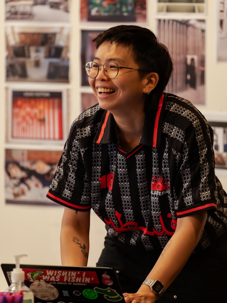 Jean Tong stands at a desk, leaning down over a laptop, looking up and laughing