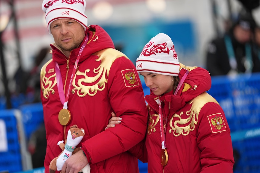 A woman holds the arm of a taller man, both dressed in red puffer jackets and red-and-white woollen caps.