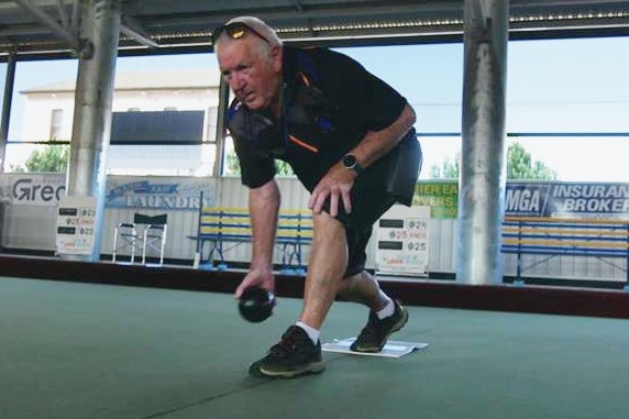 An older man wearing darker clothes rolling a ball down a bowls pitch on a shaded field on a sunny day.