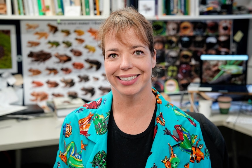 A woman wearing a bright blue frog-themed shirt smiles at the camera against a background of a frog poster