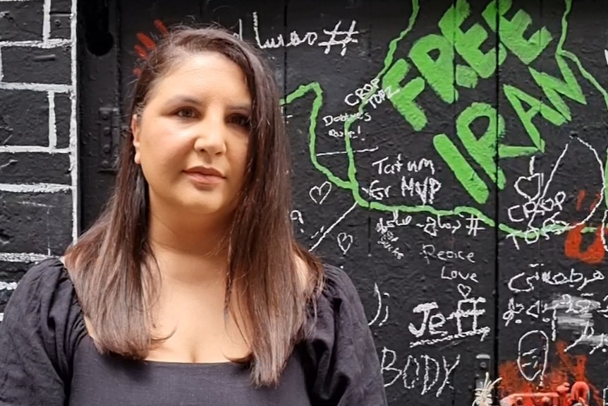A woman stands in front of a wall with grafitti saying "free Iran".   