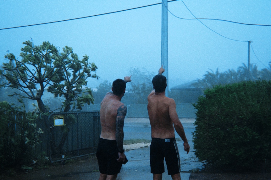 Two shirtless men point at power lines in stromy weather