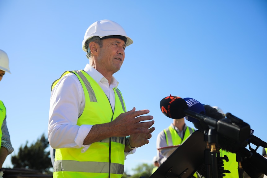 WA Premier Roger Cook speaking into microphones at a media conference outside, wearing a hi-vis vest and white hard hat.