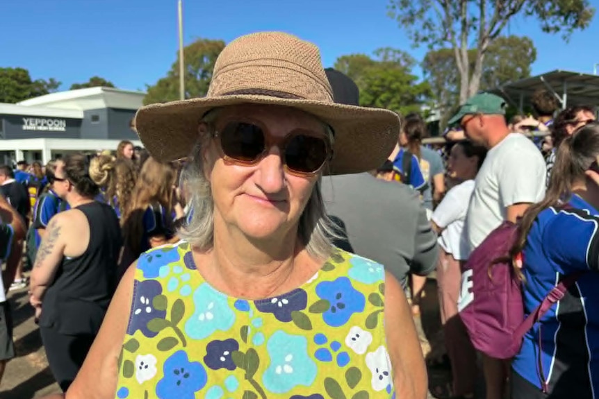 An older woman with a hat and sunglasses stands in front of a school gate.