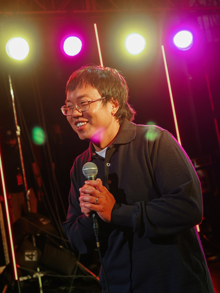 A TV still of Aaron Chen, 30, Chinese Australian, in glasses, performing on stage, smiling and clasping a mic in both hands.