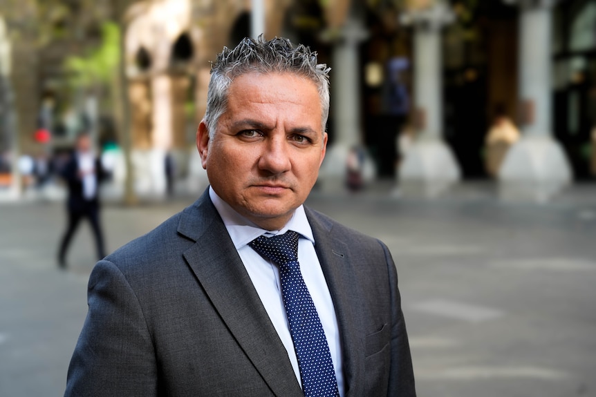 A man with short spiky salt and pepper hair in a charcoal suit and navy tie looks serious.