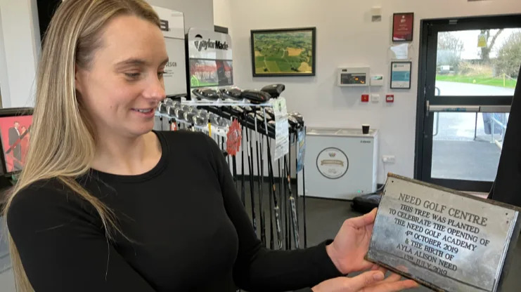 A woman with blonde hair, wearing a black top, is holding a silver plaque with the words Need Golf Centre