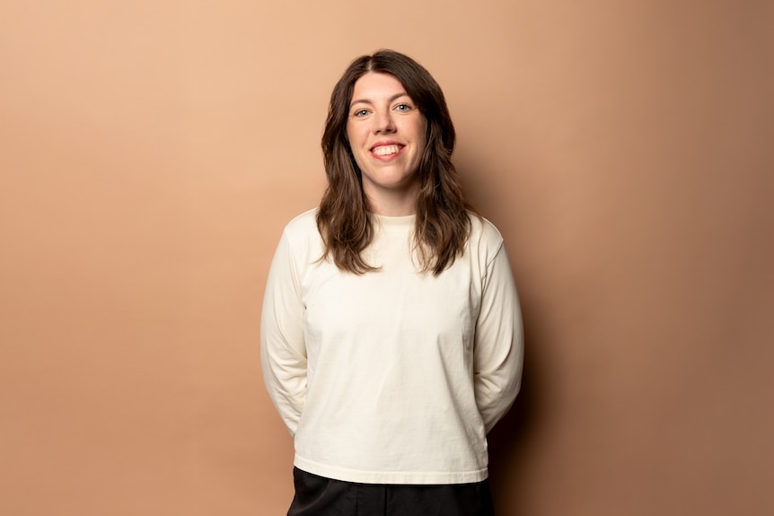 Alex Ward smiles brightly. She is wearing a white long-sleeved shirt, her arms tucked behind her back, chin tilted up.