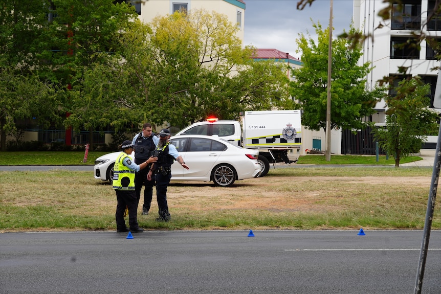 Three police officers stand talking seriously next to a road with cones on it.