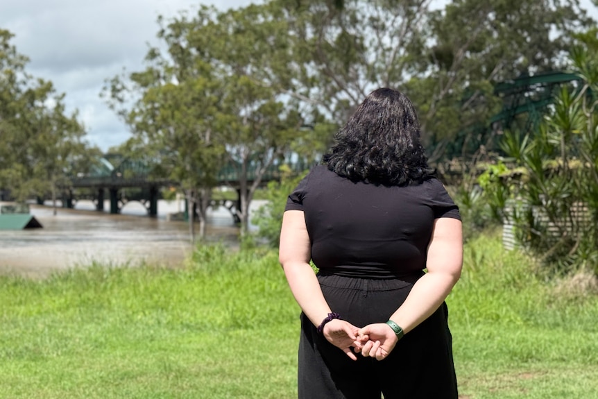 A woman faces the water with her hands clasped behind her back.