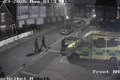 three hooded figures cross a road, walking towards an ambulance