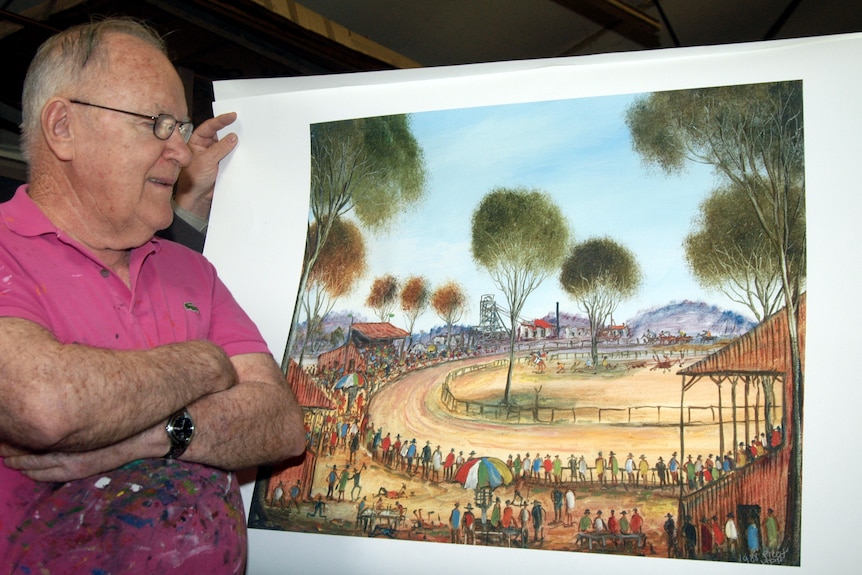 A man in a pink shirt and glasses standing with his arms crossed looking at a painting printed on a piece of paper.