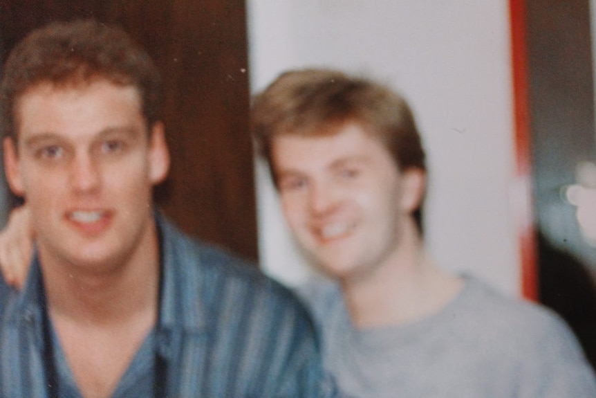 One man with high hair wearing a button up shirt and another man wearing a white t-shirt.
