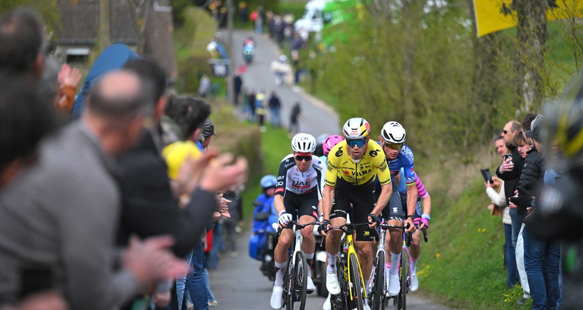 Riders approach on a narrow road with spectators on either side of the road