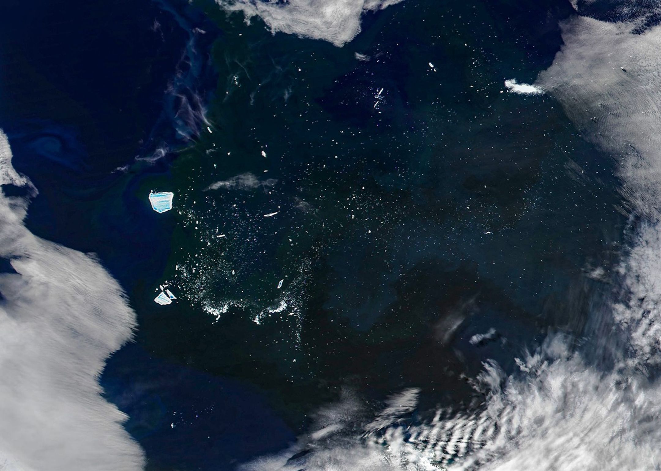 Iceberg A-23A floats in dark ocean waters colored by greenish-blue swirls of phytoplankton. Light blue pools of meltwater are visible on the surface of the iceberg. Much smaller bergs are scattered across a large area east of A-23A. Clouds along the edges of the image frame the scene.