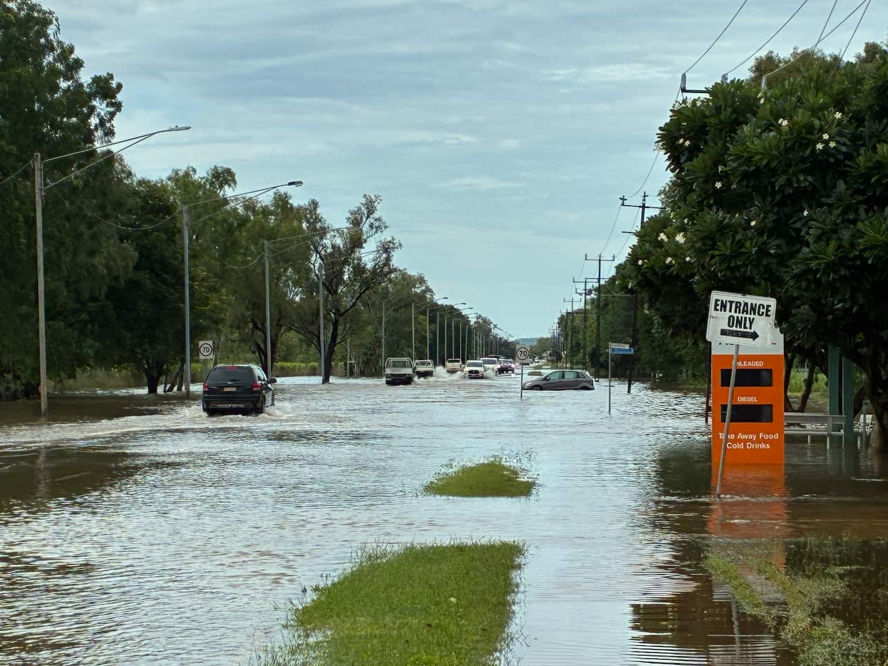 A photo of a flooded main street in Katherine. Cars are still able to pass through the water, though it appears one small car has become stranded in a deeper patch. A nearby petrol price tower sits half submerged. 