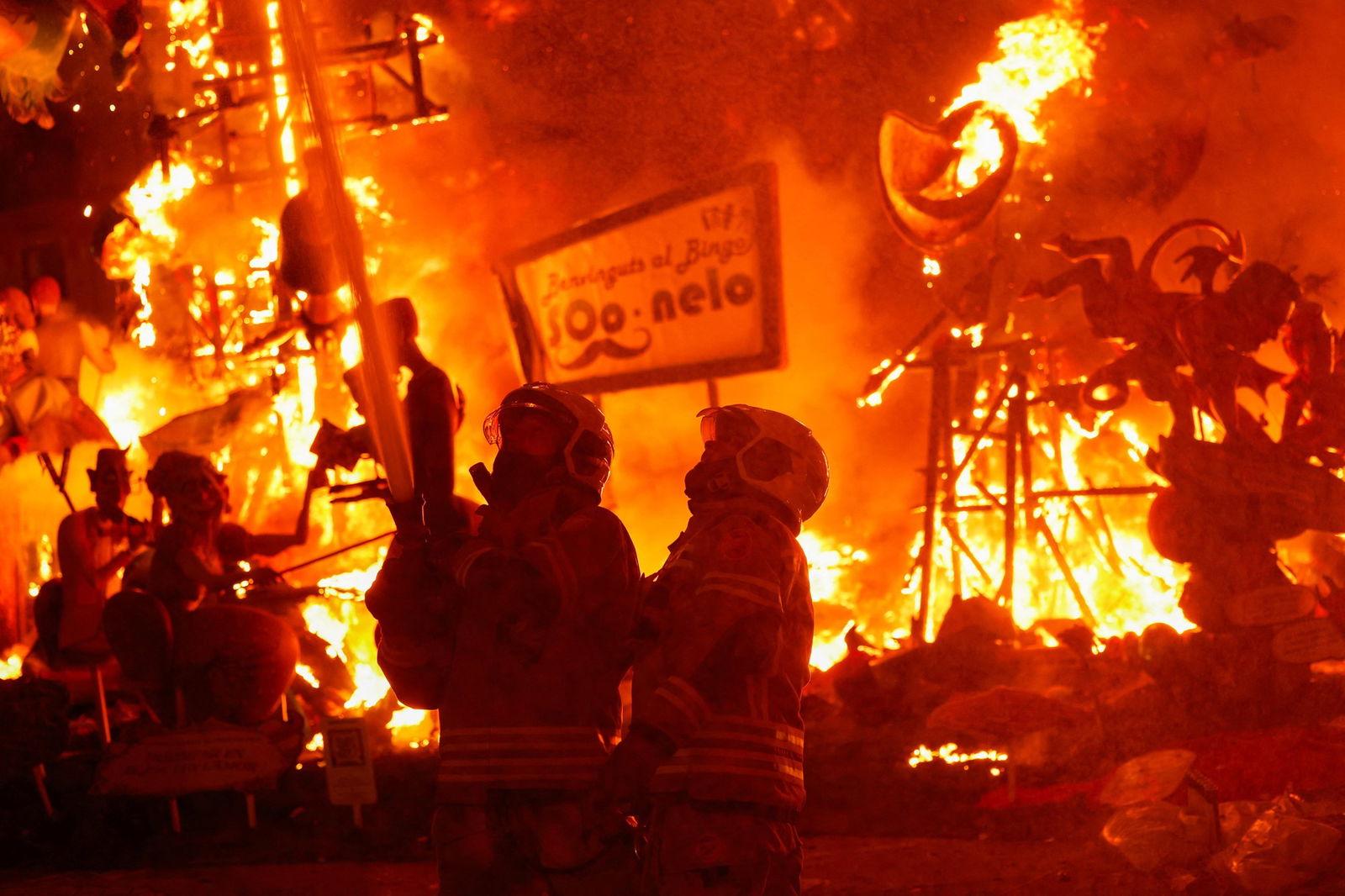 Two firefighters spray water from a large hose as they're surrounding by burning sculptures.
