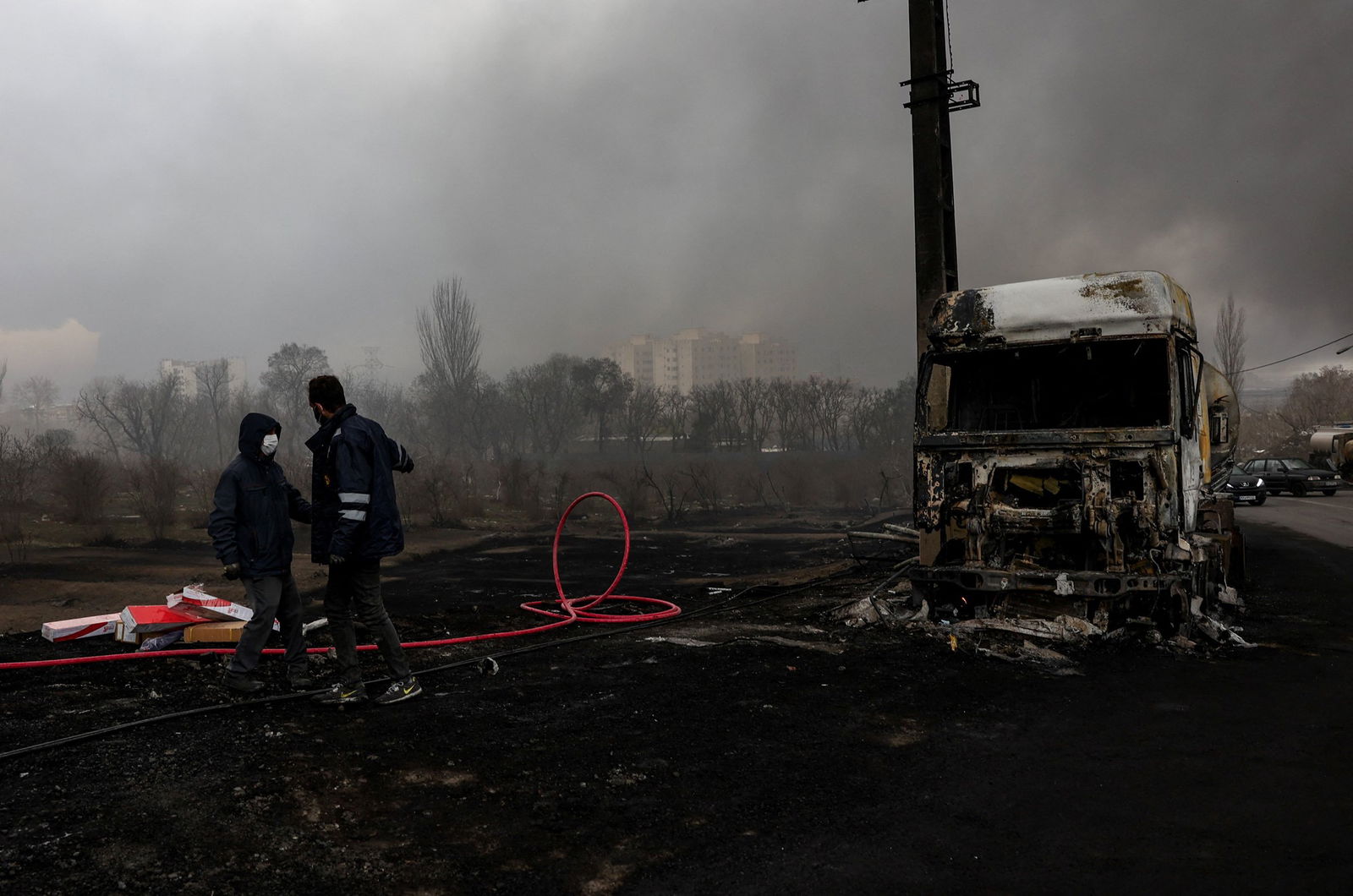Two people stand on blackened ground with a burned out truck nearby, smoke rises in the background. 