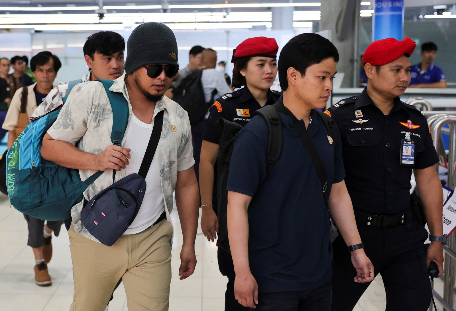 One man wearing a beanie and sunglasses with another wearing a dark blue polo. Two Thai border agents walk behind them wearing red berets. 