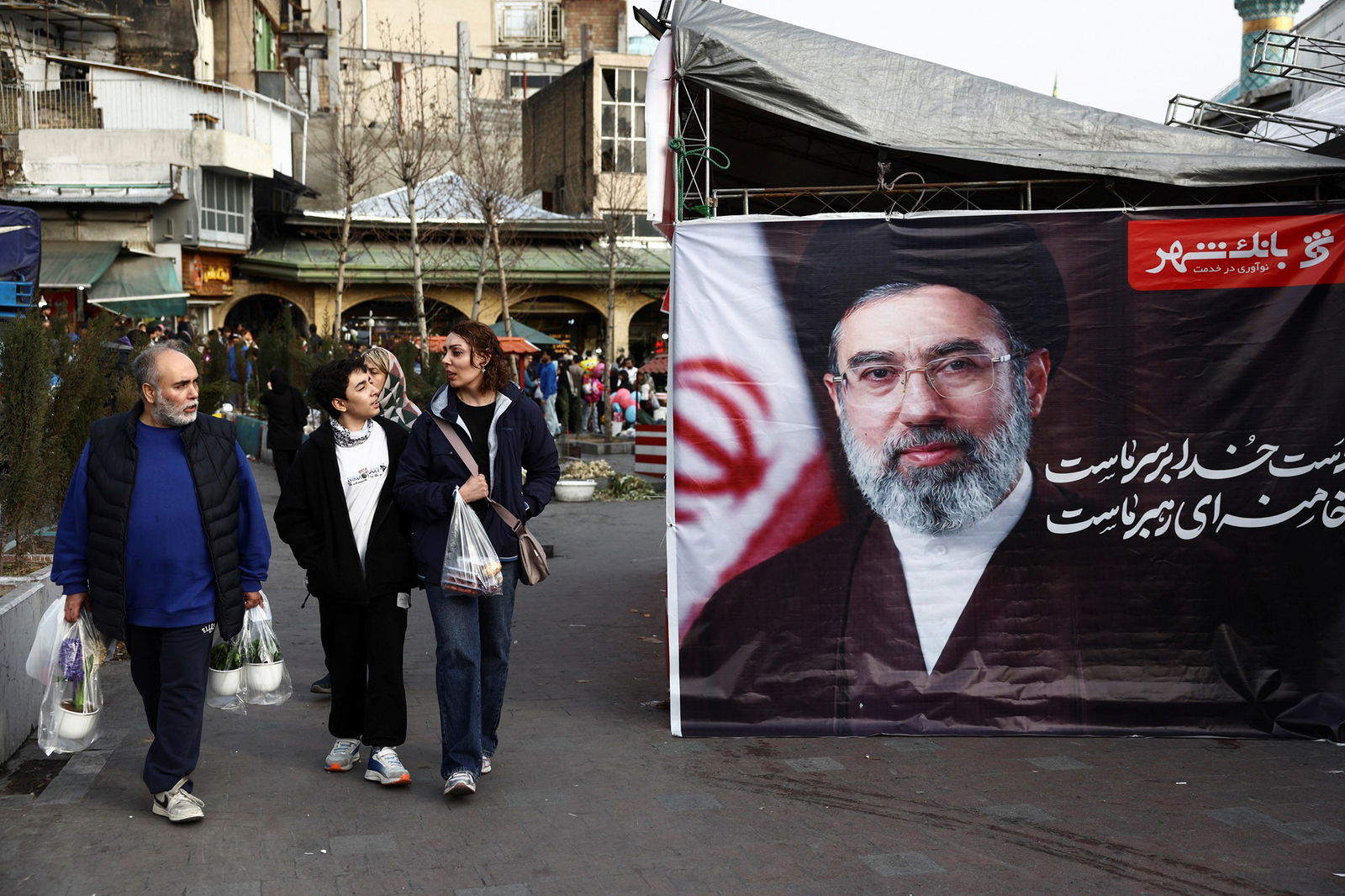 Men and women in sneakers walk past a large photo of a bearded man in a turban at an outdoor market.