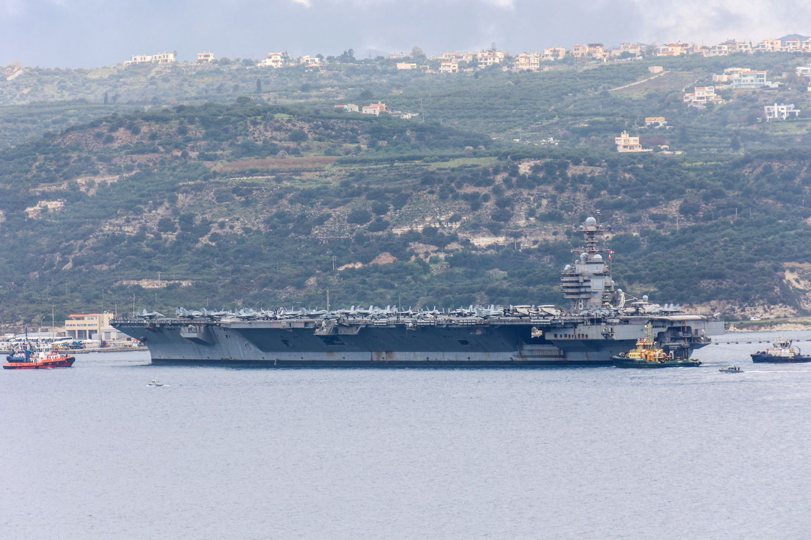 A large aircraft carrier surrounded by small boats is docked at sea off an island. 
