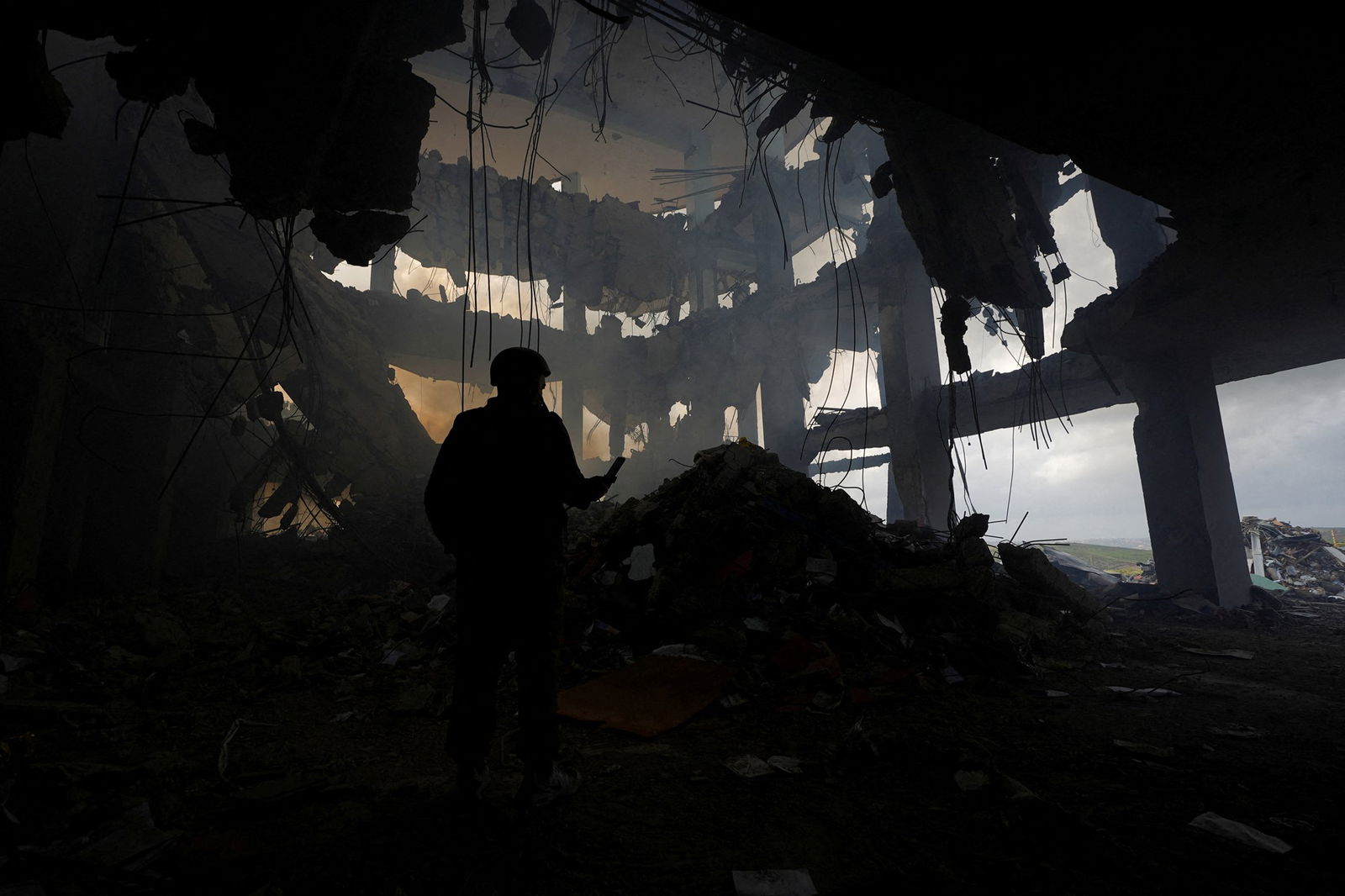 The silhouette of a person can be seen, standing in the burned out wreckage of a tall, concrete building. They stand in rubble. 
