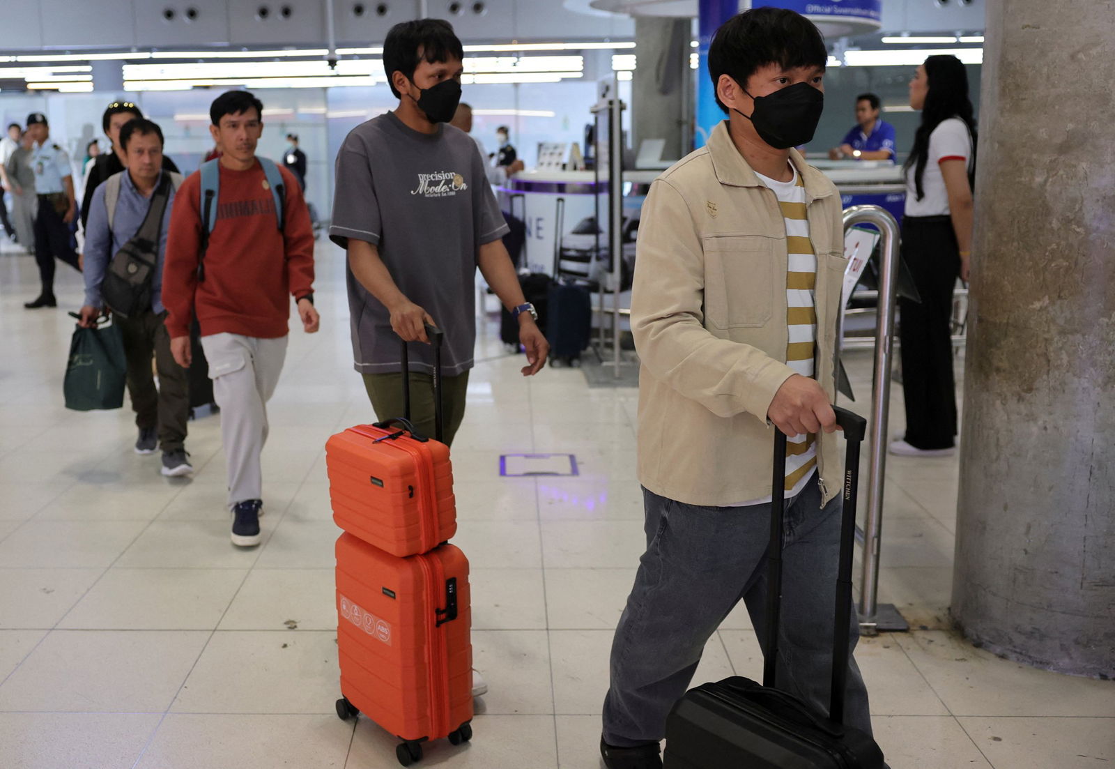 Two Thai men with their suitcases, wearing masks followed by a line of others. 