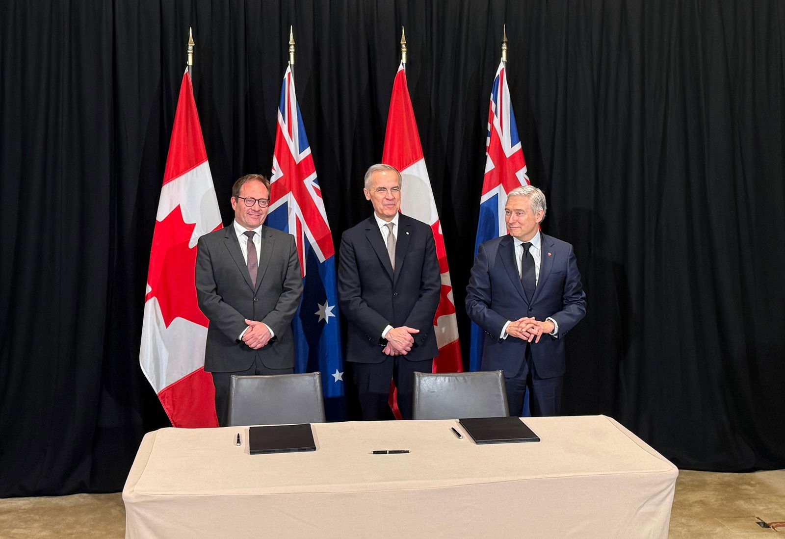 Three Caucasian men standing behind a desk in front of the Canadian and Australian flags.