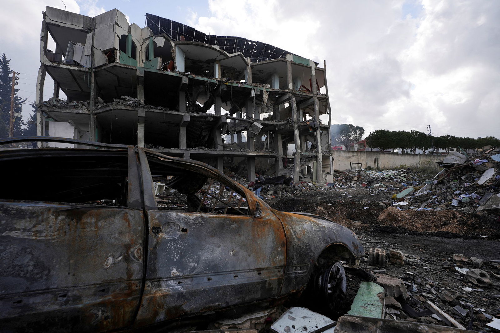 There's a rusted out shell of a care in the foreground and a destroyed four-storey building in the background. All windows and walls have been destroyed, leaving a crumbling, concrete skeleton. 
