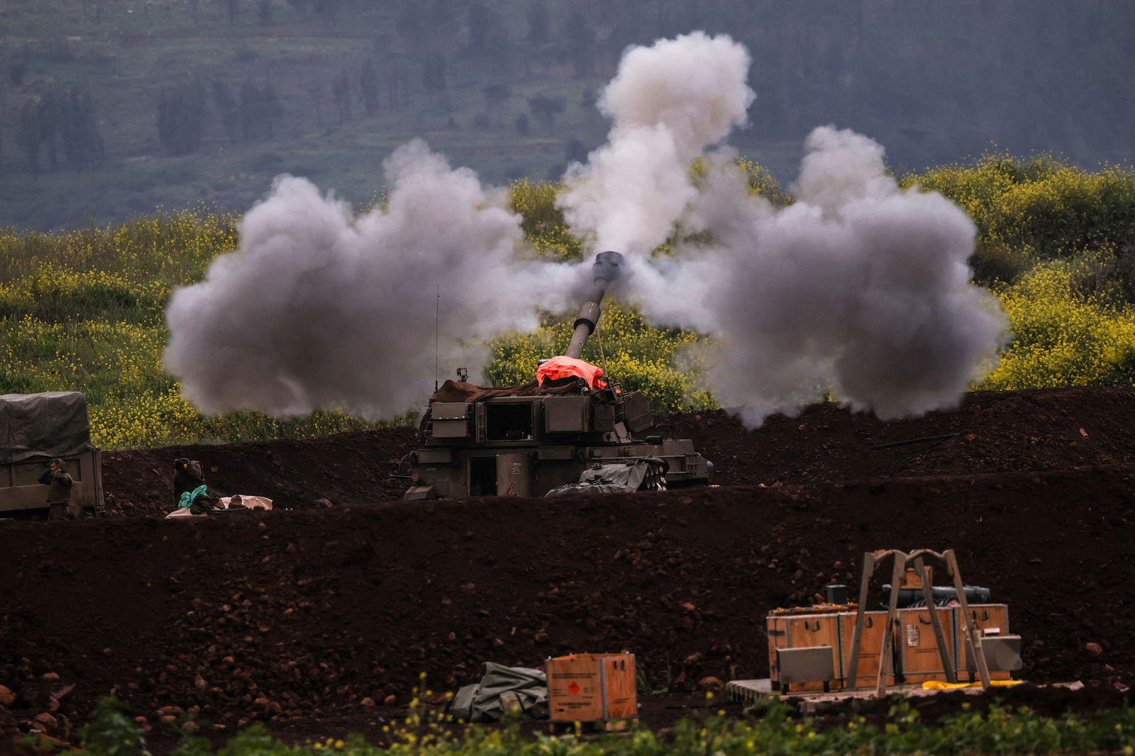 An artillery piece sits in a dirt hole behind a grassy hill. Huge plumes of smoke are shooting out of the main gun as it fires a shell. Crates of supplies are scatted in the foreground. 