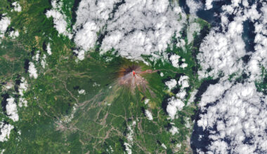 The upper slopes of Mayon volcano appear brown, with several narrow channels radiating from the crater. A red infrared heat signature appears near the summit, with red streaks extending east and southeast. The lower slopes are green and forested. Farmland and towns are visible in the lower part of the image.