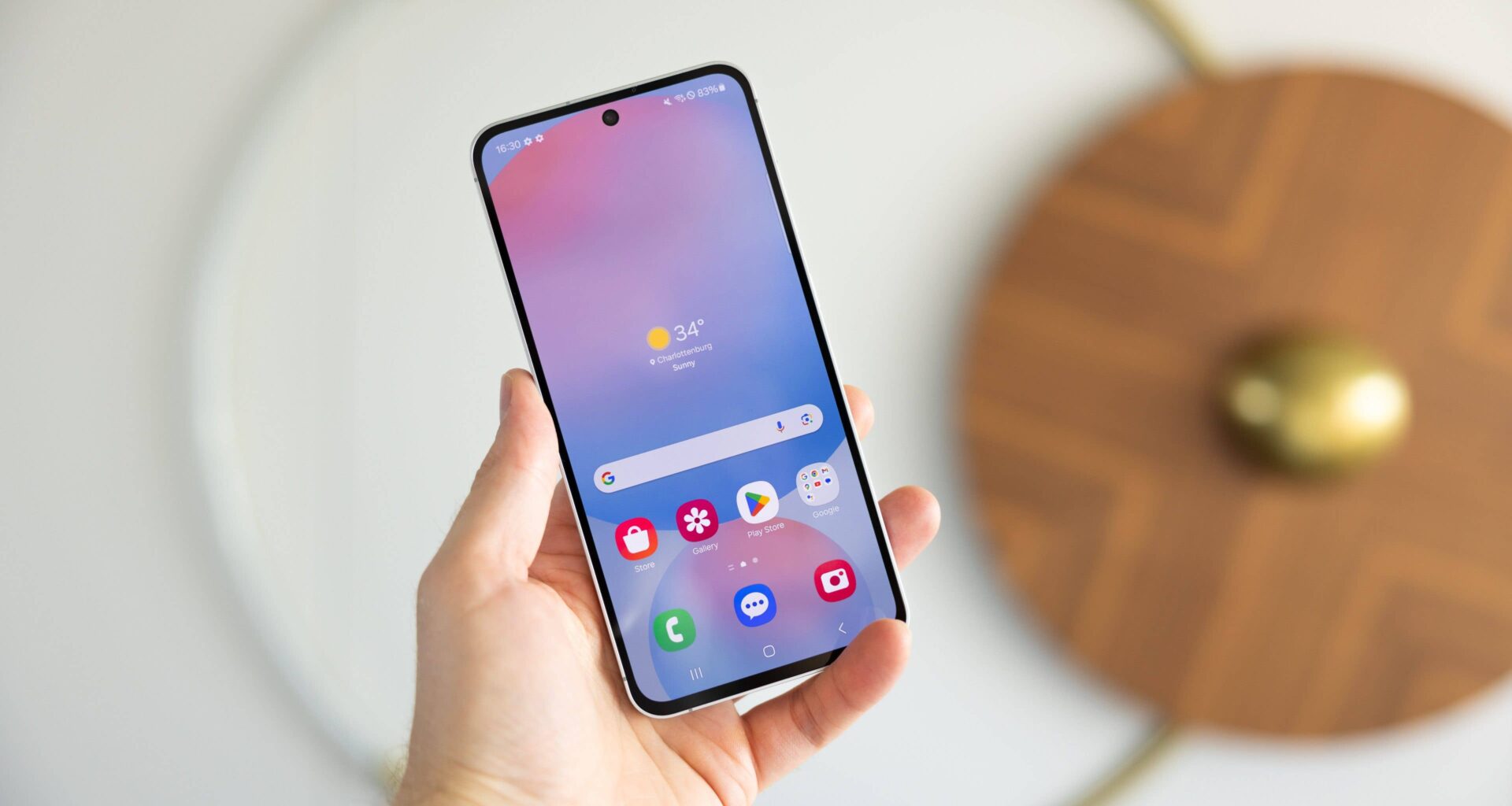 A person holding a Samsung Galaxy S24 FE smartphone displaying a weather app with a sunny 34°C.