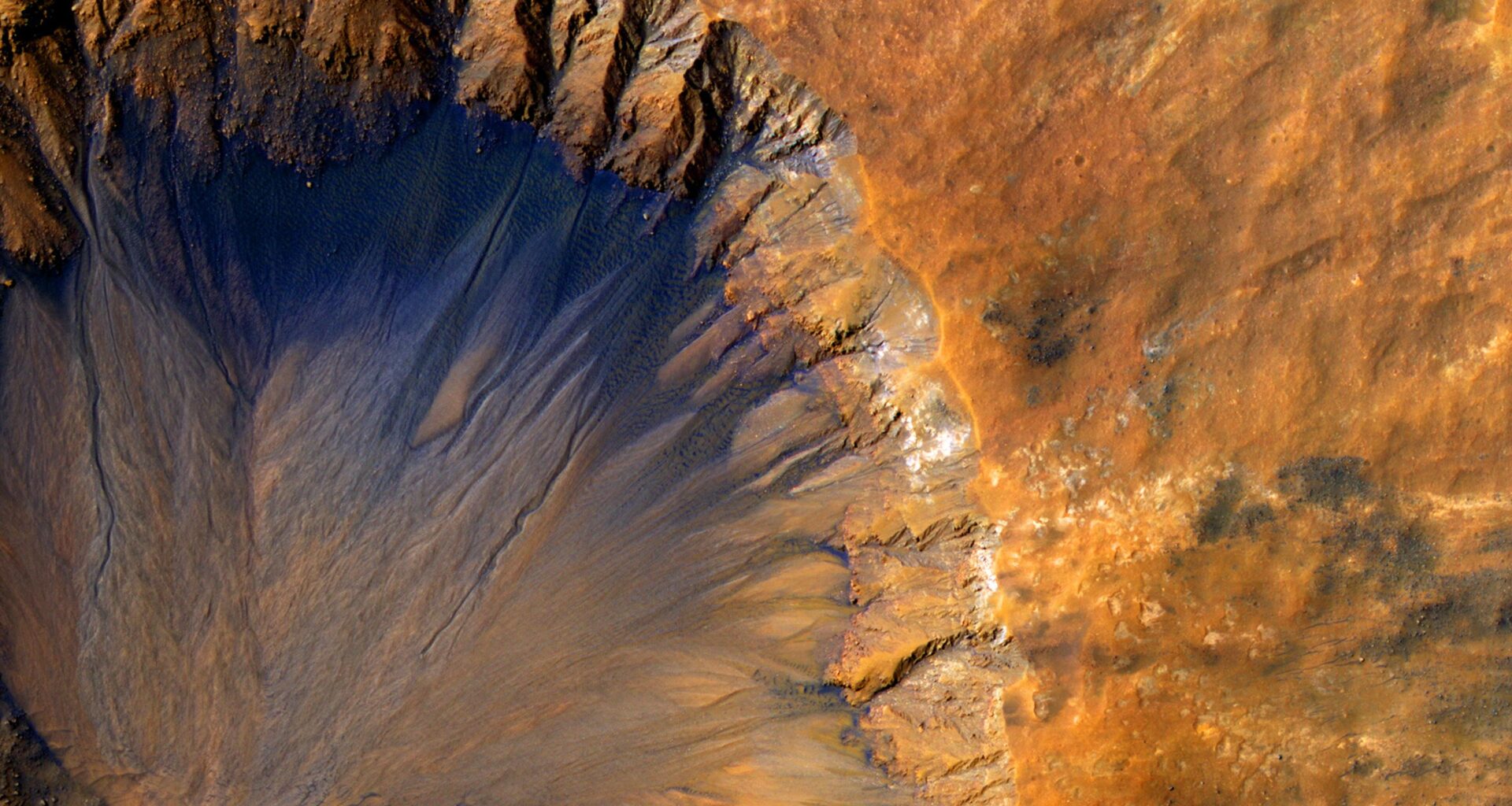 An impact crater with a sharp rim seen from above. Most of the landscape is orange, while the steep inner slope on the north side of the crater is a deep blue-black.
