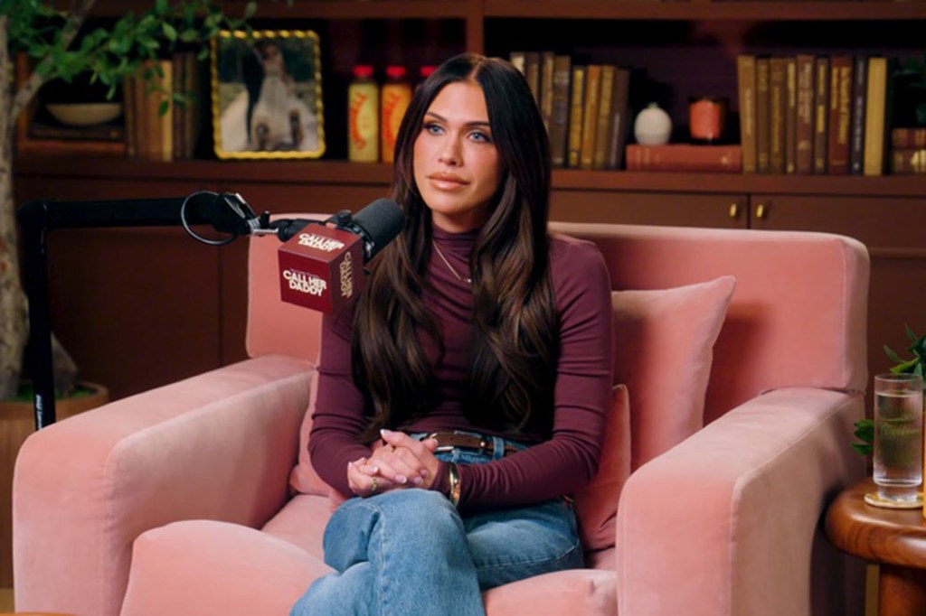 A woman sits in a pink armchair in a podcast studio, speaking into a microphone labeled “Call Her Daddy,” with bookshelves and decor in the background.