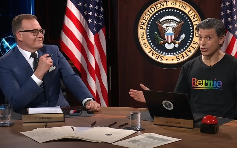 John Barron and Chas Licciardello host a political discussion at a table with the Presidential Seal, featuring Australian and American flags in the background. Chas is wearing a colourful "Bernie" t-shirt, while John appears engaged and thoughtful. Documents and a glass of water are visible on the table, highlighting the serious nature of their conversation.