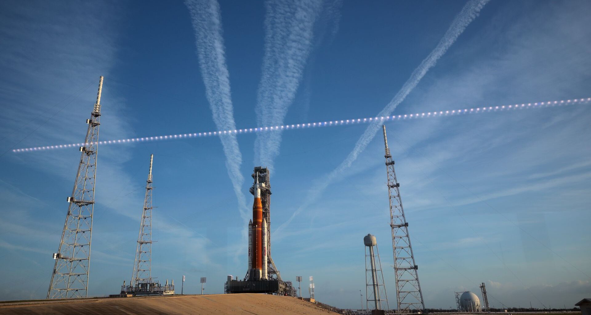 A large orange and white rocket sits on a launch pad with streaks of white clouds behind it