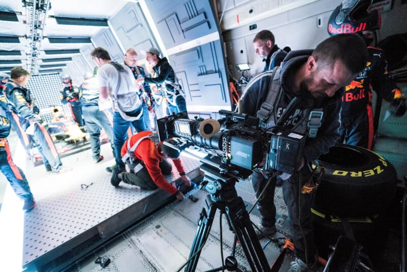 A film crew sets up a camera and equipment on a sci-fi set, with several people working in the background. Some are in harnesses, and racing suits with Red Bull logos are visible. The scene is brightly lit.