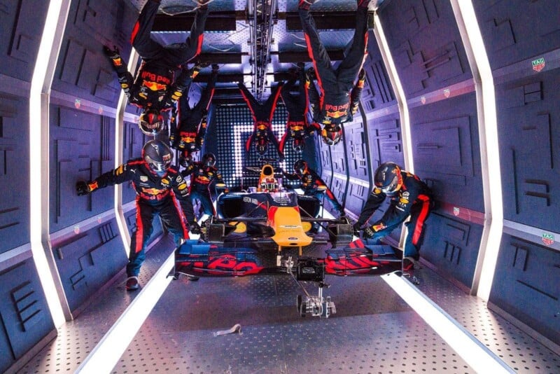 A Formula 1 pit crew, dressed in Red Bull uniforms, performs a pit stop on a race car inside a futuristic tunnel, with some crew members upside down as if they are experiencing zero gravity.