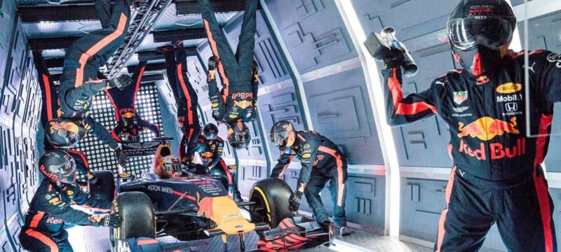 A Formula 1 pit crew in Red Bull uniforms performs a pit stop on a race car inside a futuristic, metallic garage. Some crew members appear to be working upside down, suggesting zero gravity.
