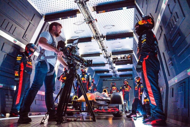 A film crew member operates a camera in a futuristic garage set, surrounded by people in Red Bull racing suits and helmets, with a Formula 1 car in the center under bright, artificial lighting.