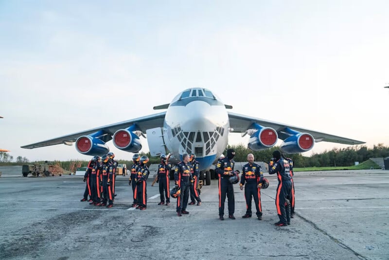 A group of people in coordinated jumpsuits stands in front of a large jet aircraft on a runway, talking and holding helmets, with trees and vehicles visible in the background.