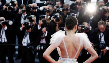 A woman in a glamorous dress poses with her back to the camera on a red carpet, facing a crowd of photographers taking pictures with bright flashes.