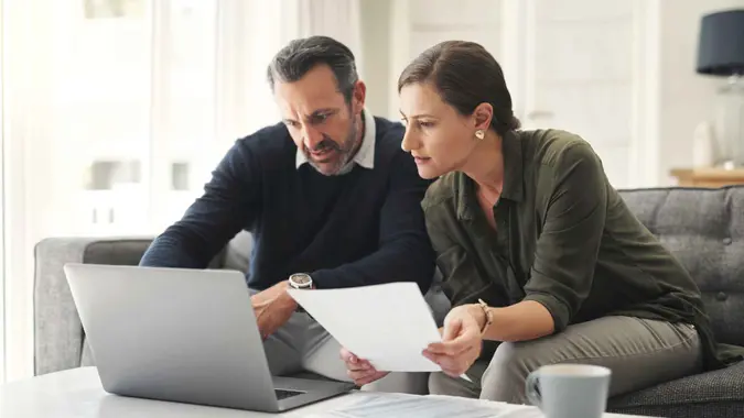 A couple reviews financial paperwork together while sitting at a table with a laptop open in front of them.