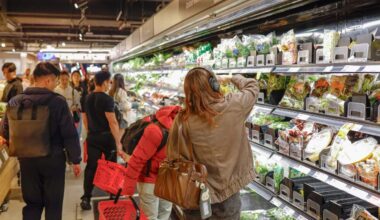 A high-angle shot shows several shoppers in a busy supermarket aisle, including a woman in a brown jacket and headphones reaching for packaged salad leaves on a refrigerated shelf.