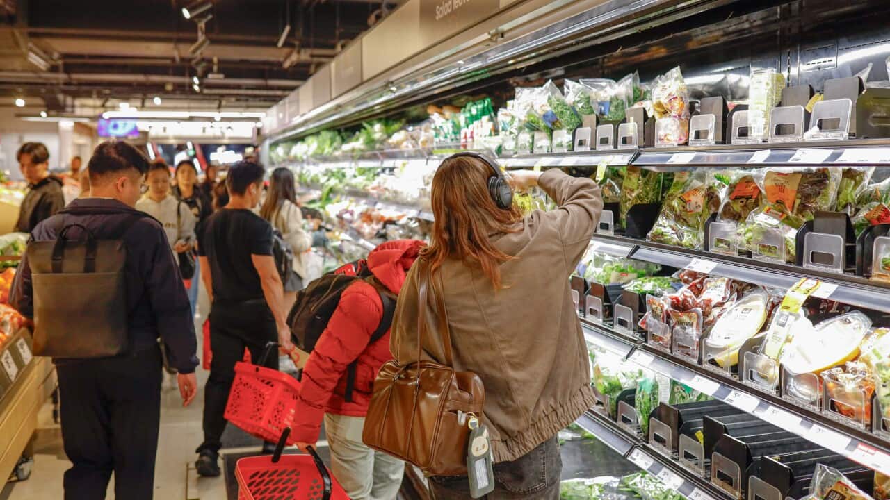 A high-angle shot shows several shoppers in a busy supermarket aisle, including a woman in a brown jacket and headphones reaching for packaged salad leaves on a refrigerated shelf.