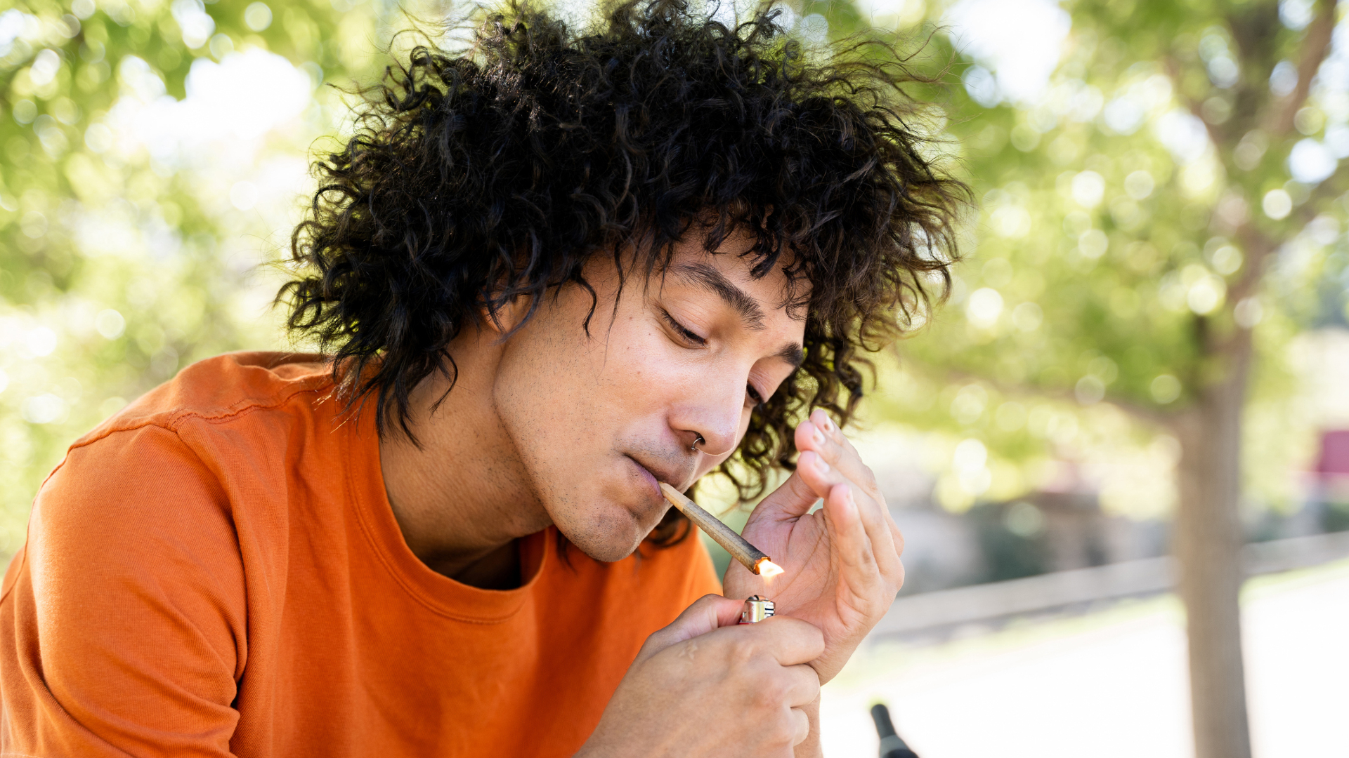 A man with long wavy black hair wearing an orange shirt lights a joint with a lighter