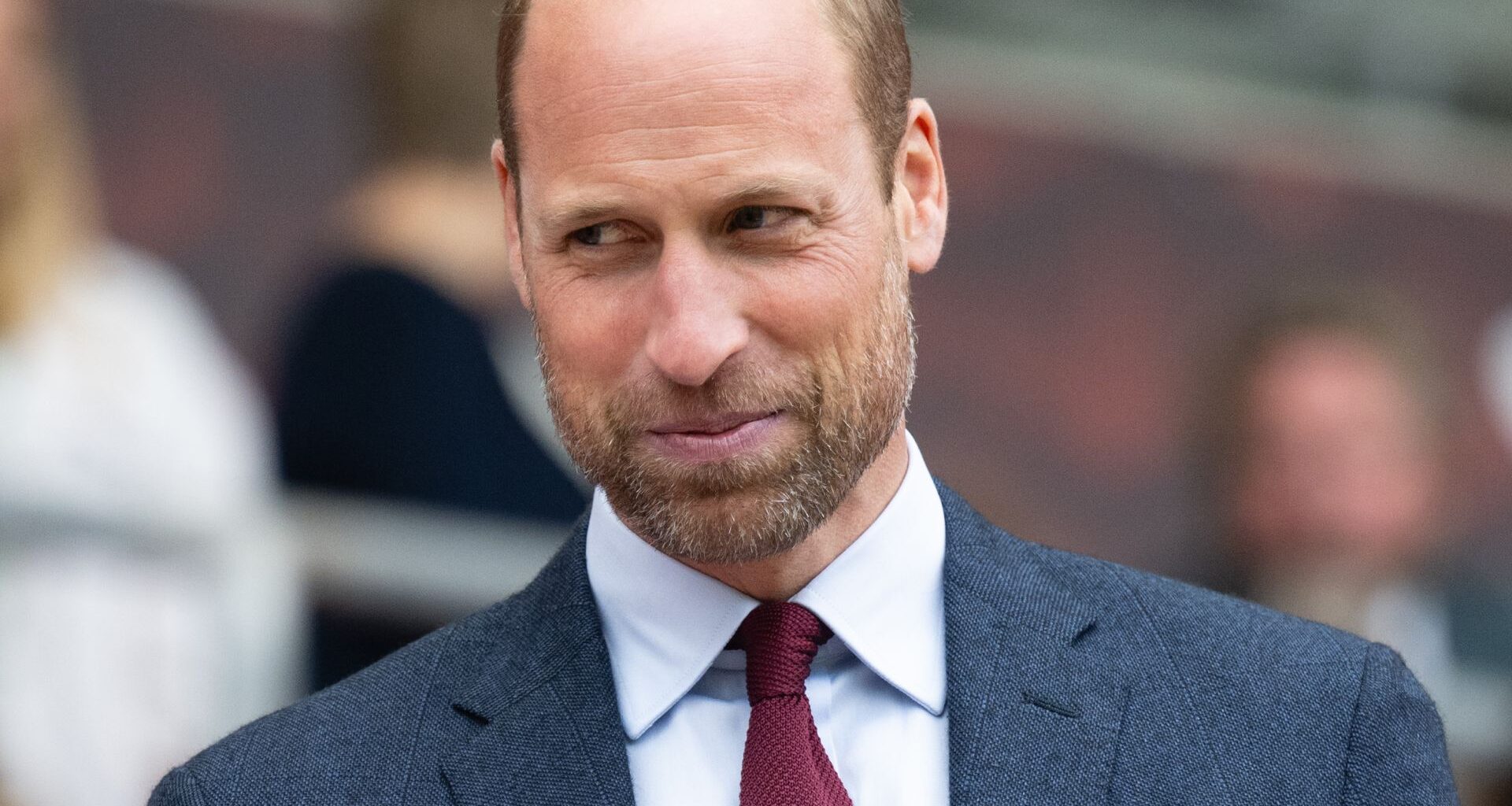 Prince William smiling while wearing a dark gray suit, a dark red tie, and a white shirt